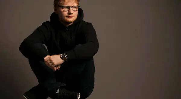 Ed Sheeran w czarnej bluzie z kapturem i okularach siedzi ze skrzyżowanymi nogami.