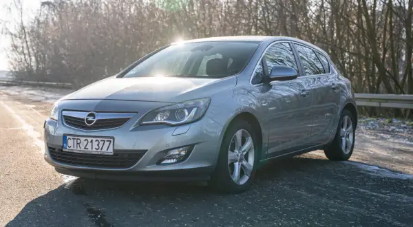 Szary Opel Astra 2011 na drodze, z drzewami w tle i słońcem odbijającym się od karoserii.