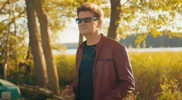 Mężczyzna w skórzanej kurtce, niczym król imperium disco polo, pozuje na tle jesiennego krajobrazu nad jeziorem.