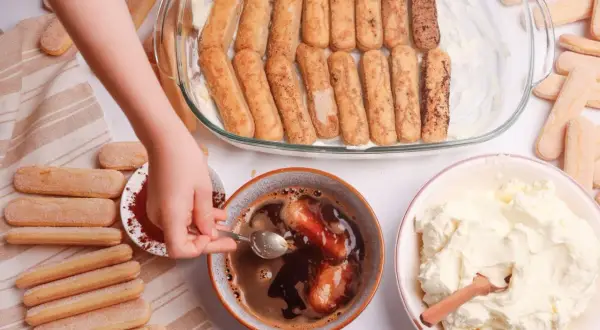 Dziecko przygotowuje tiramisu. Zanurza biszkopty w kawie, obok krem i kakao.