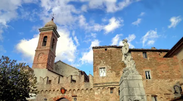 Poggio San Marcello: campanile in mattoni, un arco fortificato e una statua di bronzo sotto un cielo azzurro con nuvole bianche.