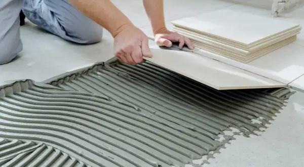 Hands carefully place a tile over cement board, with a stack of tiles nearby.