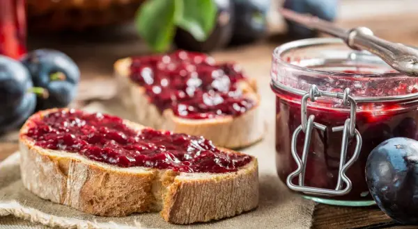 Zwei Scheiben Brot mit Zwetschgenmarmelade mit Zimt, daneben ein Glas der Marmelade und frische Zwetschgen.