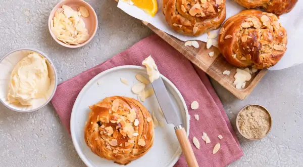 Goldbraune Kardamom-Knoten, bestreut mit Mandelblättchen, warten auf Butter und Orangenscheiben.