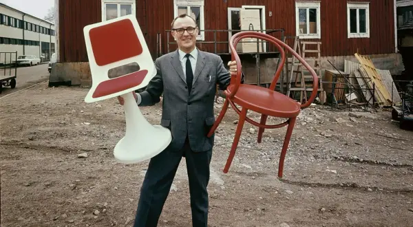 Man i kostym håller en röd stol och en vit stol med röd sits. Kanske en tidig idé för Ingvar Kamprad utbildning i möbeldesign.