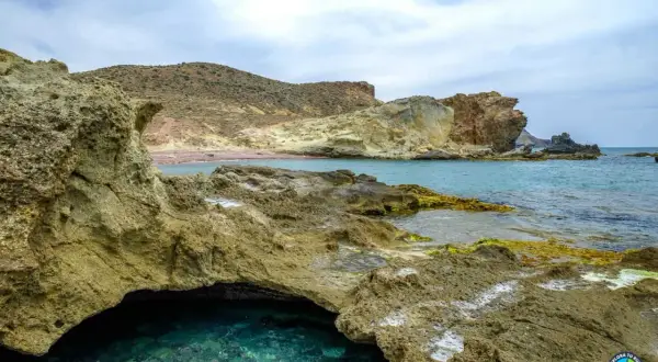 Agua turquesa en una cueva natural en la playa del carbón, con rocas volcánicas y montañas áridas al fondo.