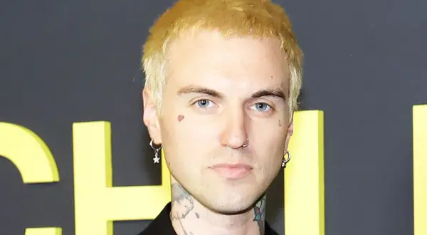 A young man with blonde hair, blue eyes, and tattoos, including "AUTOPILOT" on his neck, poses for a photo. He wears hoop earrings and a nose ring. His style is reminiscent of jordan jutes.