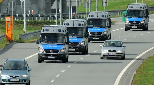 Konwój pojazdów uprzywilejowanych: policyjne furgonetki i samochód osobowy jadące autostradą.