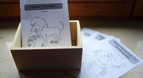 Holzkiste mit Blättern für fabeln zum lesen. Ein Blatt zeigt einen Löwen mit einem Lamm, ein anderes einen Till Eulenspiegel.