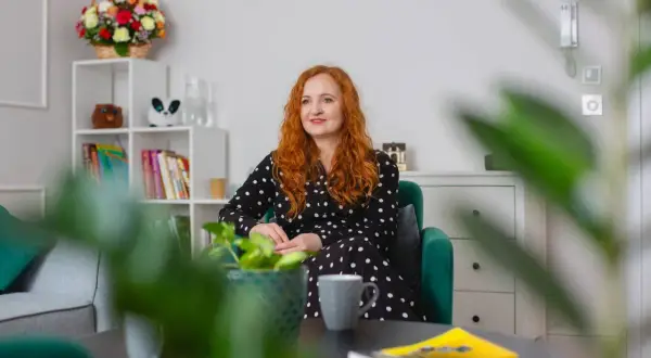 Kobieta z rudymi włosami w czarnej sukience w kropki siedzi w zielonym fotelu. Na stoliku leżą książki, a w tle widać regał z dekoracjami. To idealne miejsce na rozmowy z conners 3.