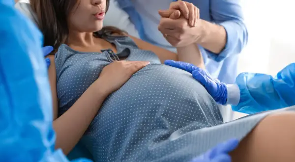 Mujer embarazada en trabajo de parto, con su pareja sosteniendo su mano y personal médico asistiendo. Se enfoca en el vientre, sugiriendo el momento de cómo provocar el parto.