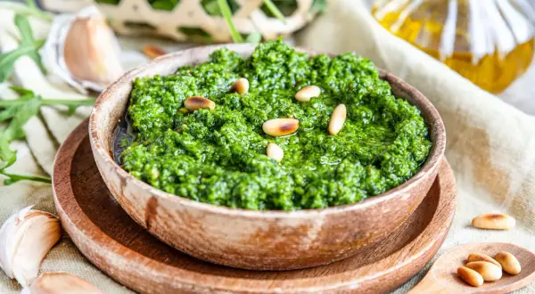 Frisches Rucola Pesto in Holzschale, garniert mit Pinienkernen. Daneben Knoblauch und Olivenöl.