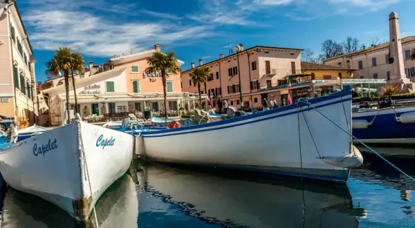 Barche a Cisano di Bardolino, con palme e case colorate sullo sfondo. Il sole splende su un cielo azzurro e limpido.