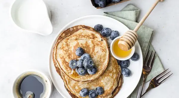 Pancake senza uova con yogurt, guarniti con mirtilli freschi e miele. Un caffè nero e una brocca di latte completano questa deliziosa colazione.