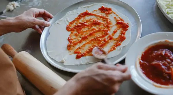 Ręce rozsmarowują pyszny sos pomidorowy do pizzy na cieście. Obok miseczka z sosem i tarta mozzarella.
