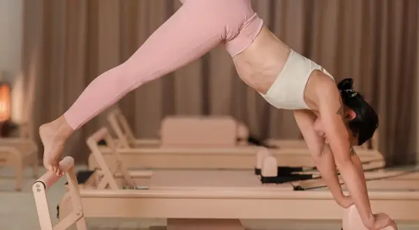 Kobieta w różowych legginsach wykonuje pozycję psa z głową w dół na reformerze. Pilates co daje? Wzmocnienie mięśni, elastyczność i harmonię.