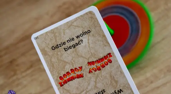 Karta z gry "Gorący ziemniak" pyta "Gdzie nie wolno biegać?". W tle kolorowy bączek.