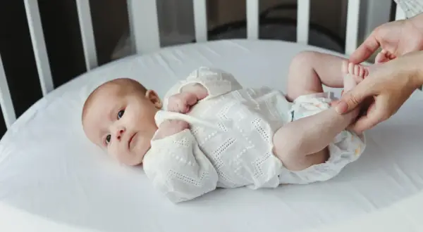 Noworodek w białej koszulce leży w łóżeczku. Dorośli delikatnie dotykają jego stópek. Tak jak widzi noworodek, świat jest rozmazany, ale pełen miłości.