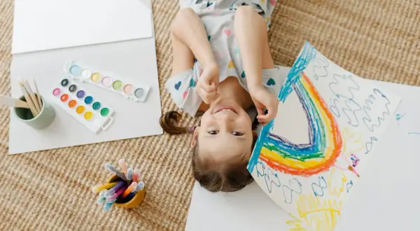 Niña sonriente haciendo un arcoíris, rodeada de acuarelas y rotuladores. Perfectas actividades de verano para niños creativos.