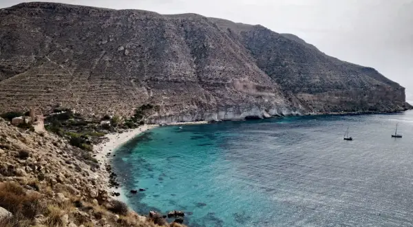 Bahía de aguas turquesas en la playa Cabo de Gata, con barcos navegando y montañas áridas al fondo.