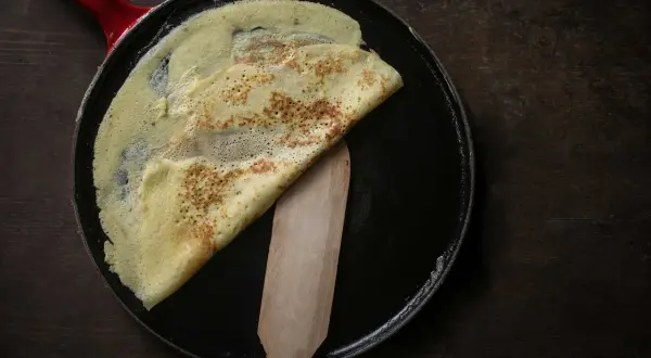 Ein goldbrauner Crêpe wird in einer Pfanne mit einem Holzspatel gefaltet. Perfekt für ein einfaches Crêpe Rezept.