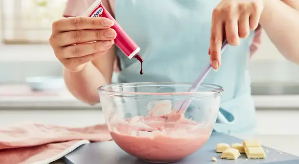 En hand droppar röd färg i en skål med rosa smet, för att skapa ett perfekt glasyr recept.