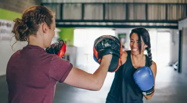Due donne si allenano nel **boxing** in palestra. Una tiene i guantoni da allenamento, l'altra indossa un guantone blu.