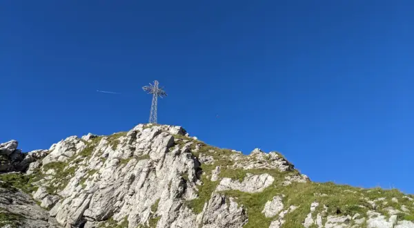 Na szczycie skalistego zbocza, porośniętego trawą, stoi metalowy krzyż. W tle błękitne niebo, a w oddali widać samolot. To widok z Giewontu, który przypomina, ile ma wysokości.