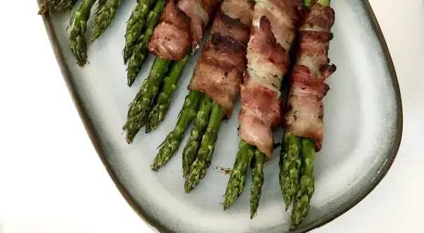 Grön sparris insvept i knaprigt bacon, serverad på en ljusblå tallrik. En utsökt kombination av grönsaker och baconlindad sparris.