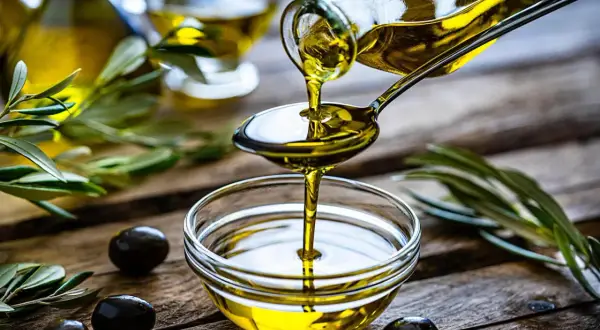 Olio di oliva non extravergine versato da un cucchiaio in una ciotola, con olive e rami d'ulivo sullo sfondo.
