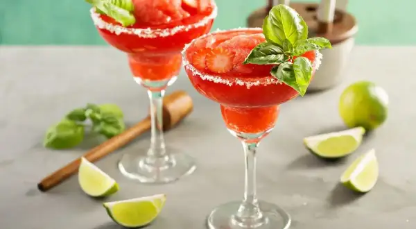 Dwa mrożone strawberry daiquiri z bazylią i cukrowym brzegiem, podane z limonkami.