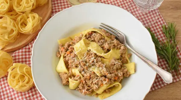 Pappardelle z bogatym ragu, które to danie, posypane parmezanem. Obok surowy makaron i gałązka rozmarynu.