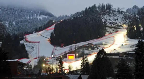 Die Skirennstrecke, beleuchtet für die Nacht, mit roten Banden und einem roten Hahnkamm-Symbol.