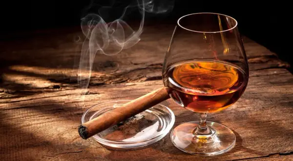 Ein Glas bernsteinfarbener Bourbon mit aufsteigendem Rauch von einer Zigarre auf einem Holztisch.