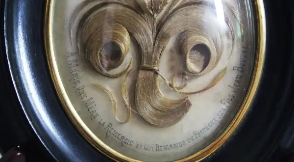 Intricate hair art in a framed oval, featuring swirling locks of blonde hair arranged like a floral bouquet. Text reads "A Marie, une mère la remercie et lui demande de protéger son fils Jean Boudet.