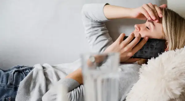 Kobieta leży na kanapie, trzymając telefon przy uchu i dotykając czoła. Wygląda na chorą, co jest dobrym powodem na zwolnienie lekarskie.