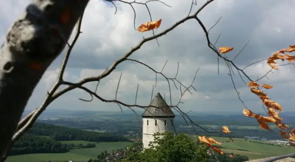 Blick durch kahle Äste auf die Burg Bielstein, ein weißer Turm mit grauem Dach ragt über grüne Hügel und eine Stadt.