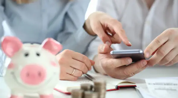 Dwie osoby planują wydatki, korzystając z telefonu i notatnika. Obok stoi skarbonka w kształcie świnki, symbolizująca oszczędności i fundusz socjalny.