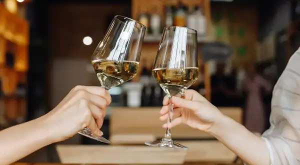 Zwei Hände halten Gläser mit einem goldenen Cuvée Wein. Ein Toast auf den Genuss in gemütlicher Atmosphäre.