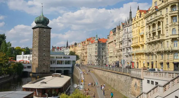 Vue de Prague que voir : tour de pierre avec dôme vert, maisons colorées le long de la rivière, et une péniche.