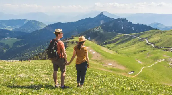 Couple en chapeau admirant un paysage de montagne verdoyant, parsemé de fleurs jaunes. Une rando Chartreuse insolite, avec des sentiers sinueux et des sommets bleutés à l'horizon.