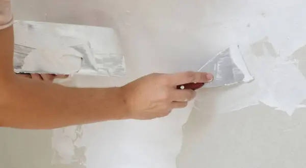 Manos aplicando masilla blanca en una pared con espátulas. La mejor masilla para paredes deja un acabado liso y perfecto.