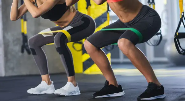 Dwie kobiety wykonują ćwiczenia z gumami oporowymi na udach, przypominające przysiady. Nie jest to hack squat maszyna, ale ćwiczenie angażujące podobne partie mięśni.