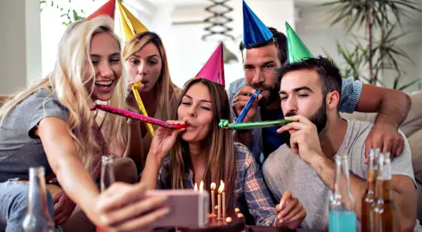 Grupa przyjaciół świętuje 70 urodziny, dmuchając w trąbki i robiąc selfie przy torcie.