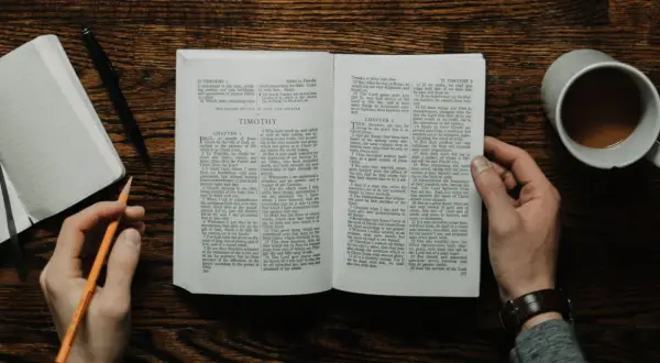 Dłonie trzymają otwartą księgę, jedną z apostolskich ksiąg Nowego Testamentu, obok leży notatnik i kubek kawy.