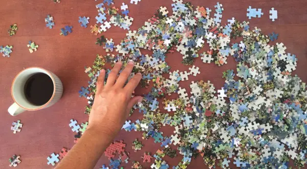 Ręka układa puzzle do układania na drewnianym stole obok kubka kawy.