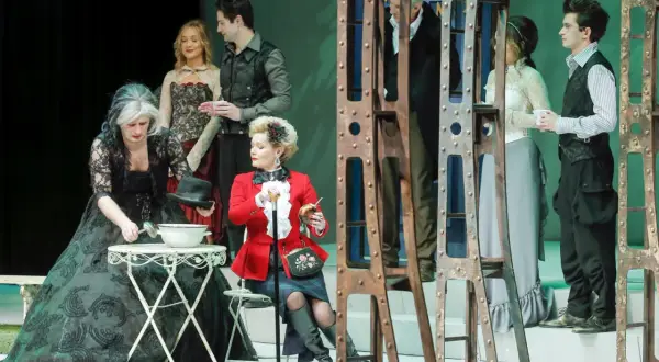 Scena z przedstawienia "Teatr kwadrat za jakie grzechy". Kobieta w czarnej sukni nalewa coś z misy, obok siedzi dama w czerwonym żakiecie.