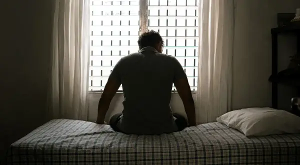 Hombre sentado en la cama, mirando por la ventana. Parece sumido en sus pensamientos, atravesando una **noche oscura del alma**.
