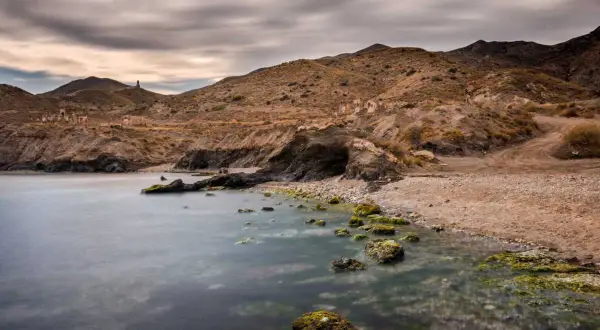 Mar tranquilo en la cala invencible Almería, con rocas cubiertas de algas y ruinas en la ladera.