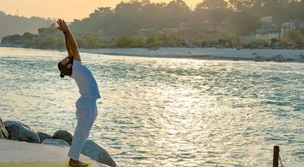 Hombre practica yoga Sivananda en la orilla de un río al amanecer, con los brazos extendidos hacia el cielo.